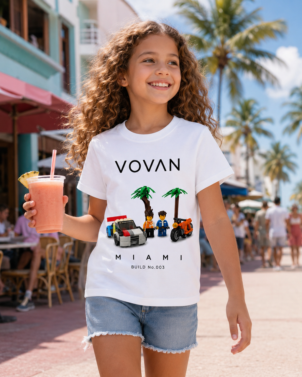 Girl wearing a white t-shirt with 'VOVAN Miami' build no.003 design by 9 years old boy champion LEGO, holding a smoothie in an outdoor setting.